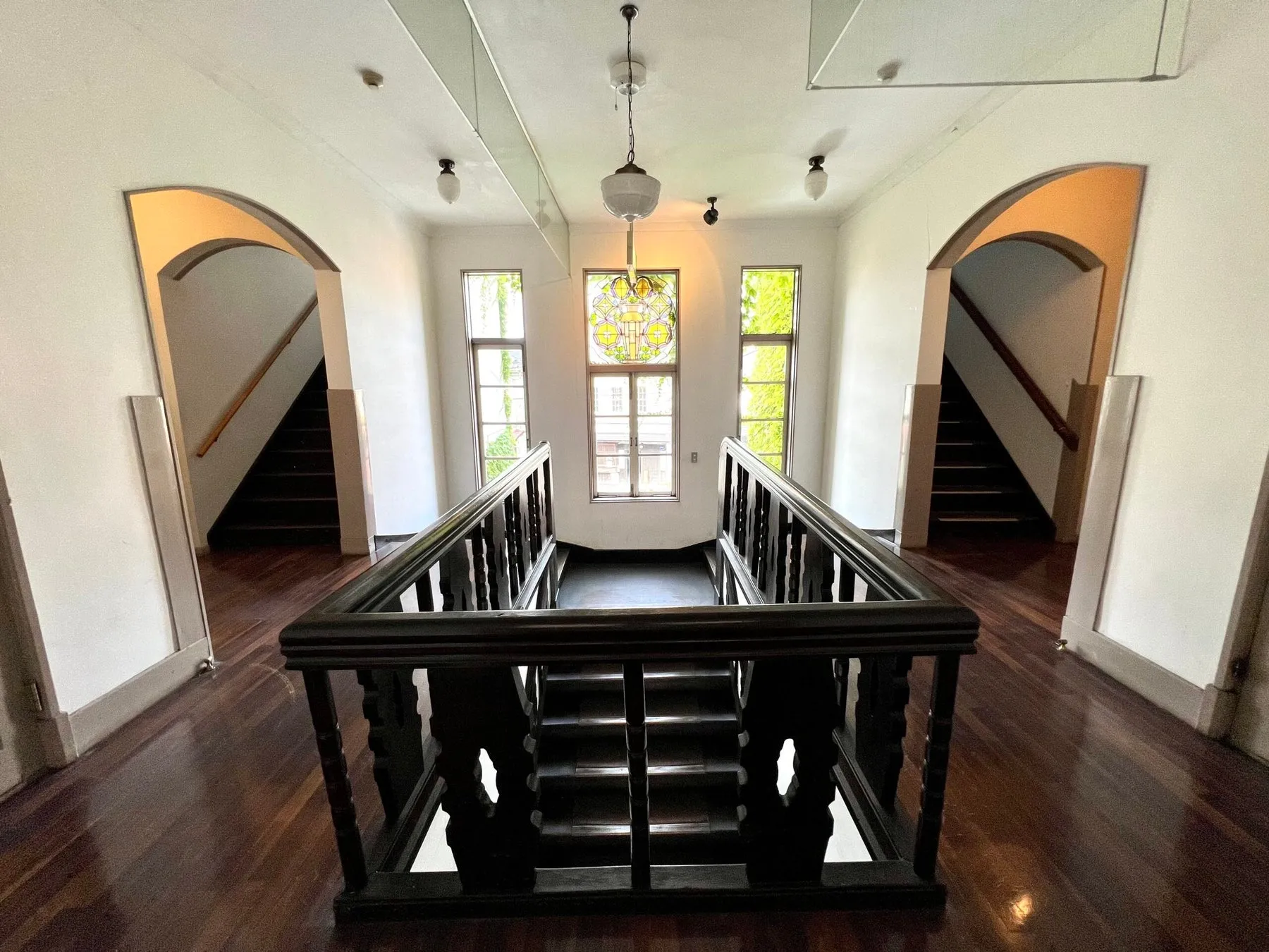 歴史的建造物のホールまたは踊り場を捉えた写真。中央にはステンドグラスの窓があり、両側には木製の手すりを持つ階段が対称的に配置されています。