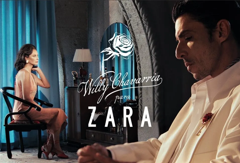 男性と女性のファッション広告。女性は椅子に座り、男性は画面の右側に立っています。背景には、鏡、ランプ、装飾的なオブジェクトがあります。