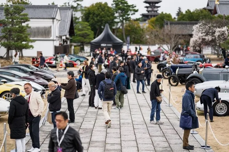 クラシックカーのイベントの様子。多くの人が集まり、様々な種類のクラシックカーが展示されている。背景には日本の寺院のような建物が見える。