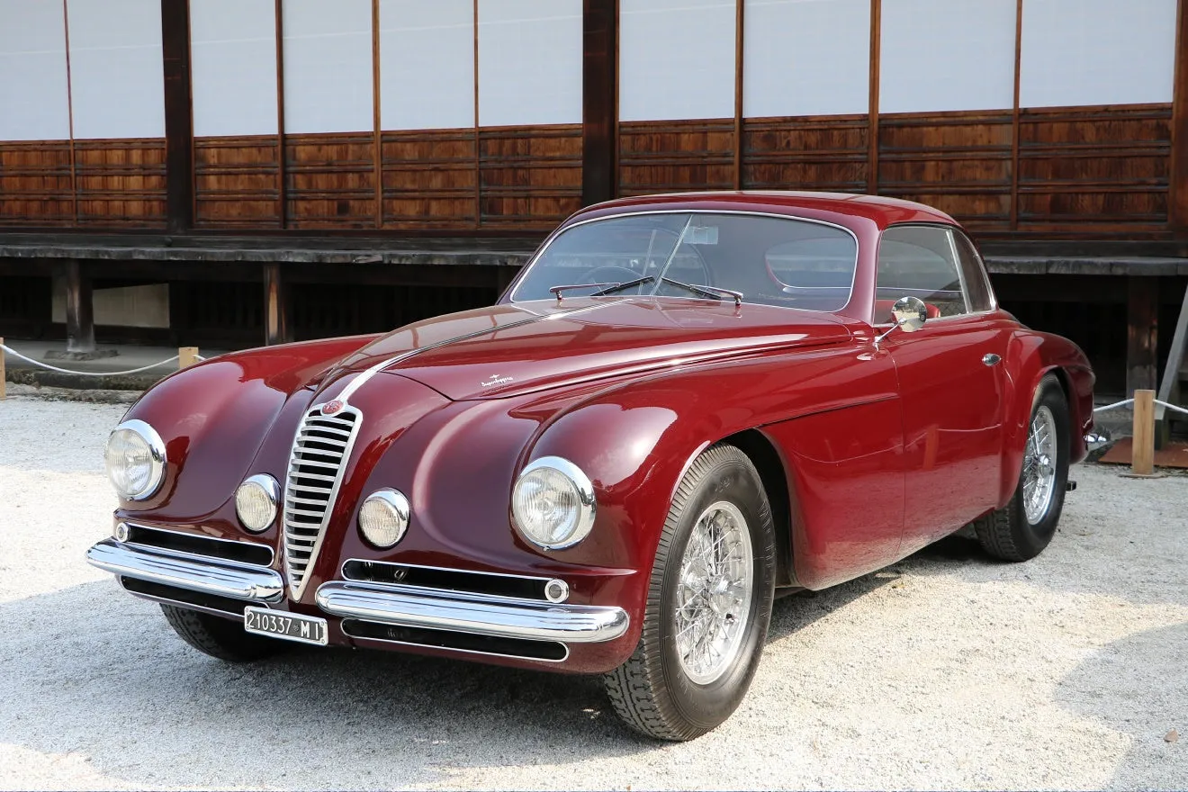 鮮やかな赤色のクラシックカーが、日本の伝統的な建築物を背景に写っています。車の流れるような曲線と、背景の木造建築のコントラストが印象的です。