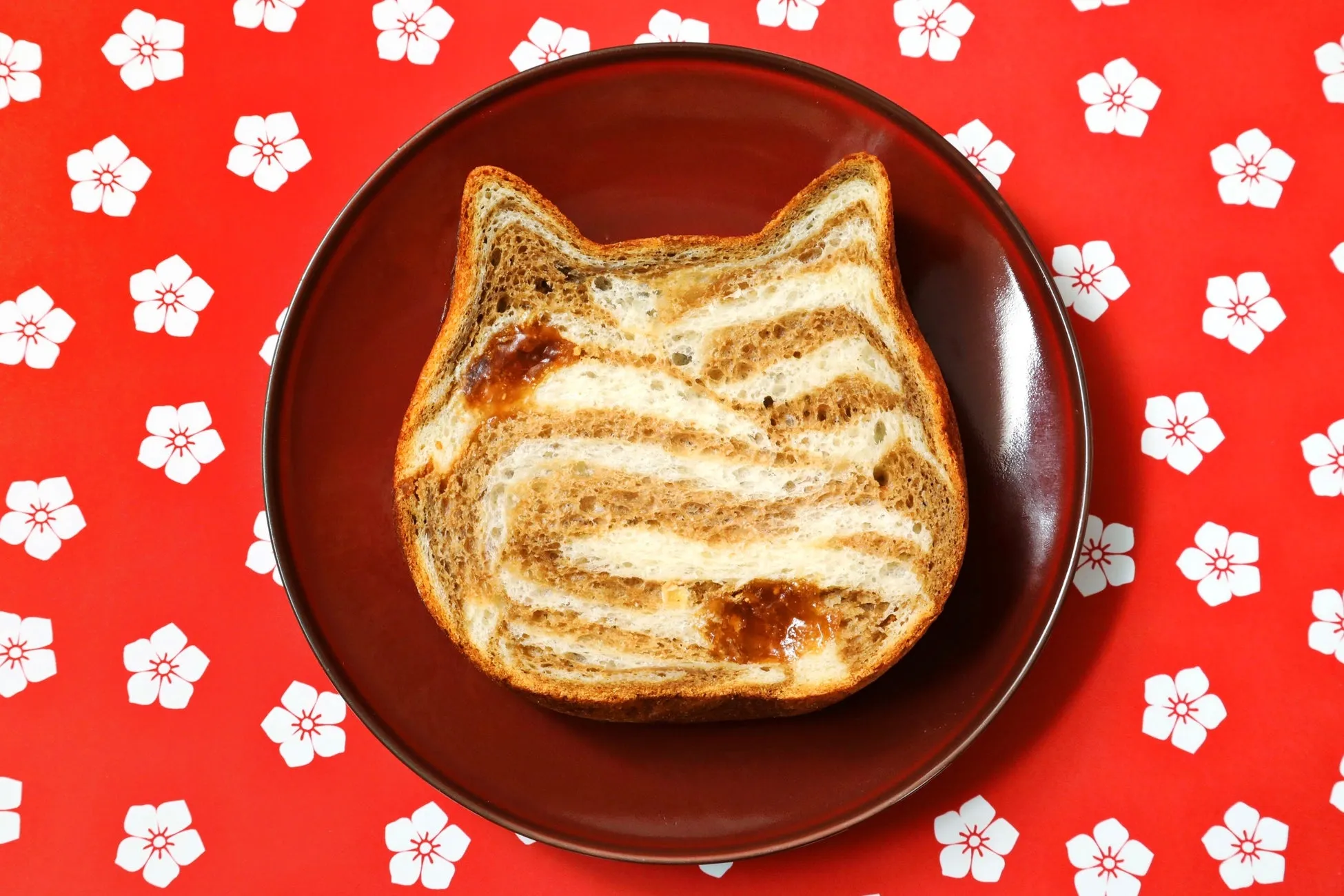 猫の形をしたトーストが皿の上に置かれており、背景は赤い花柄のデザインになっています。