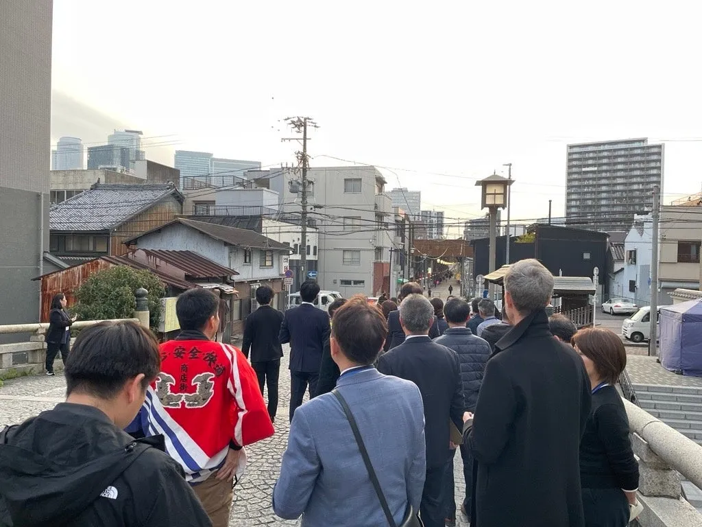 多くの人が橋の上を歩いている様子を捉えた写真。手前には赤いジャケットを着た人がおり、背中には漢字が書かれています。背景には古い建物や高層ビルが混在し、日本の街並みが写っています。