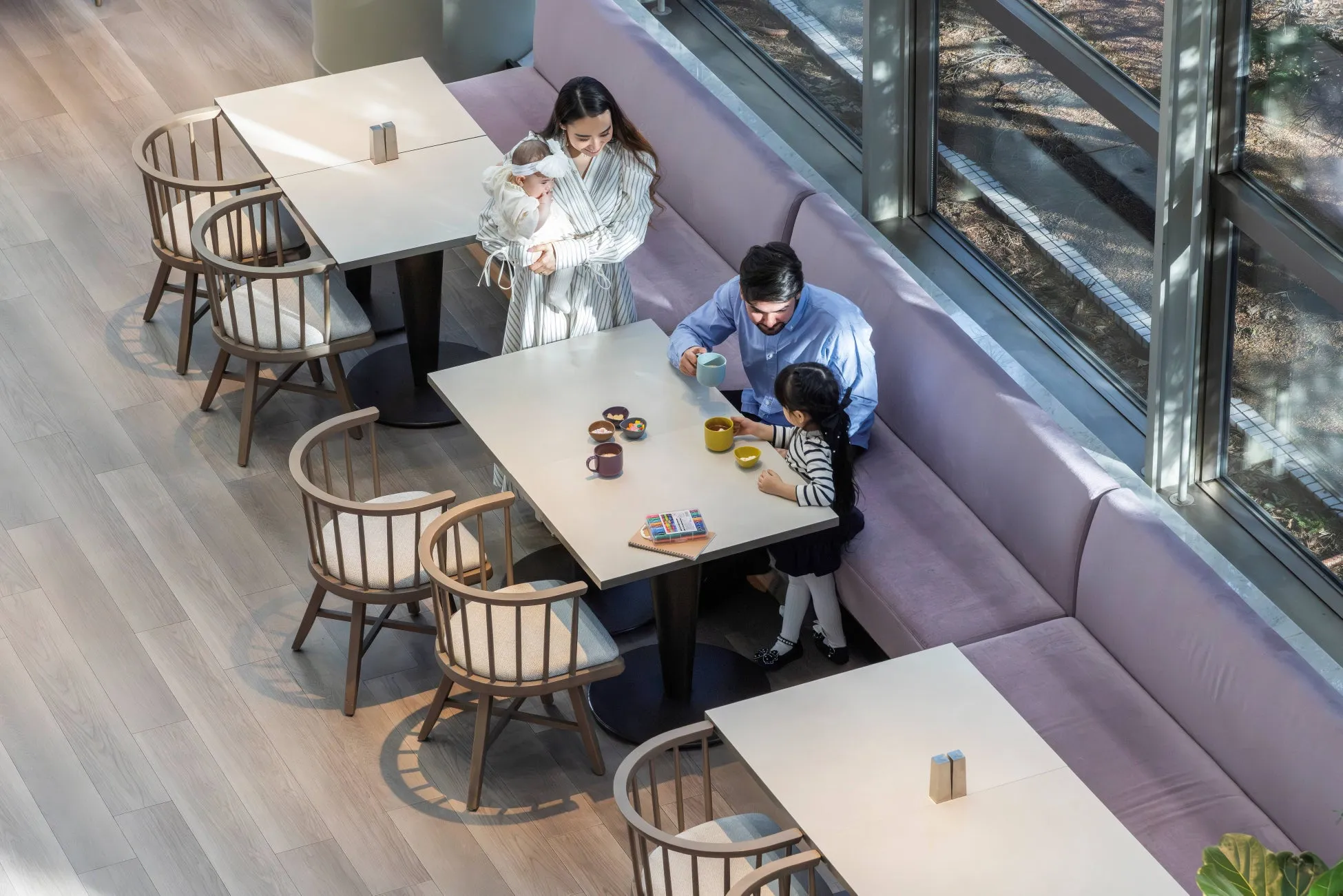 カフェで家族がくつろいでいる様子を捉えた写真です。母親は赤ちゃんを抱き、父親と幼い子供はテーブルを囲んで飲み物を楽しんでいます。モダンなデザインのテーブルと椅子が配置され、窓から自然光が差し込む明るい空間です。