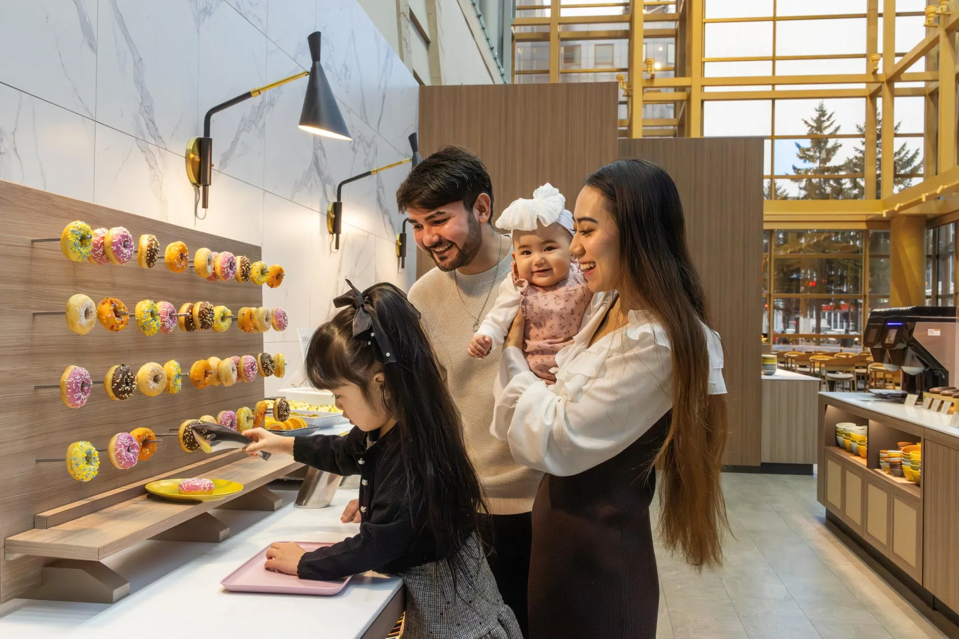 ドーナツビュッフェの前で家族が楽しそうにしている様子。ドーナツを選ぶ女の子、赤ちゃんを抱く母親、それを見守る父親の姿が写っています。