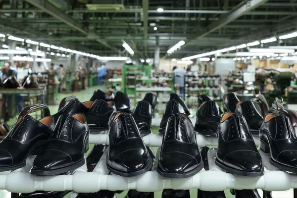 靴工場で並べられた黒い革靴のクローズアップ写真。背景には工場の機械と作業員が写っています。