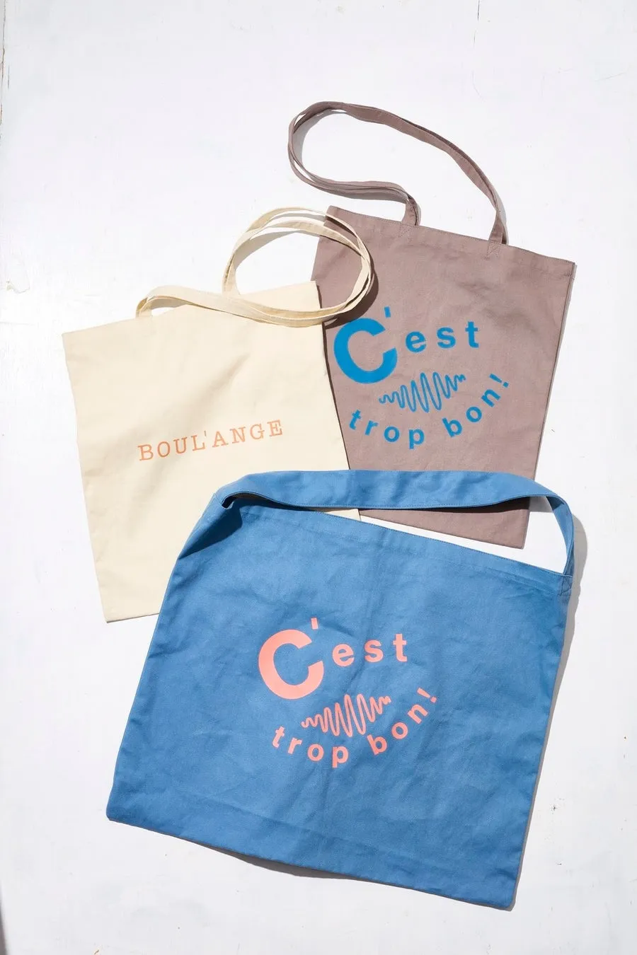 3つのトートバッグが写っている写真。生成りのバッグには「BOUL'ANGE」の文字、茶色のバッグと青色のバッグには「C'est trop bon!」の文字がプリントされている。