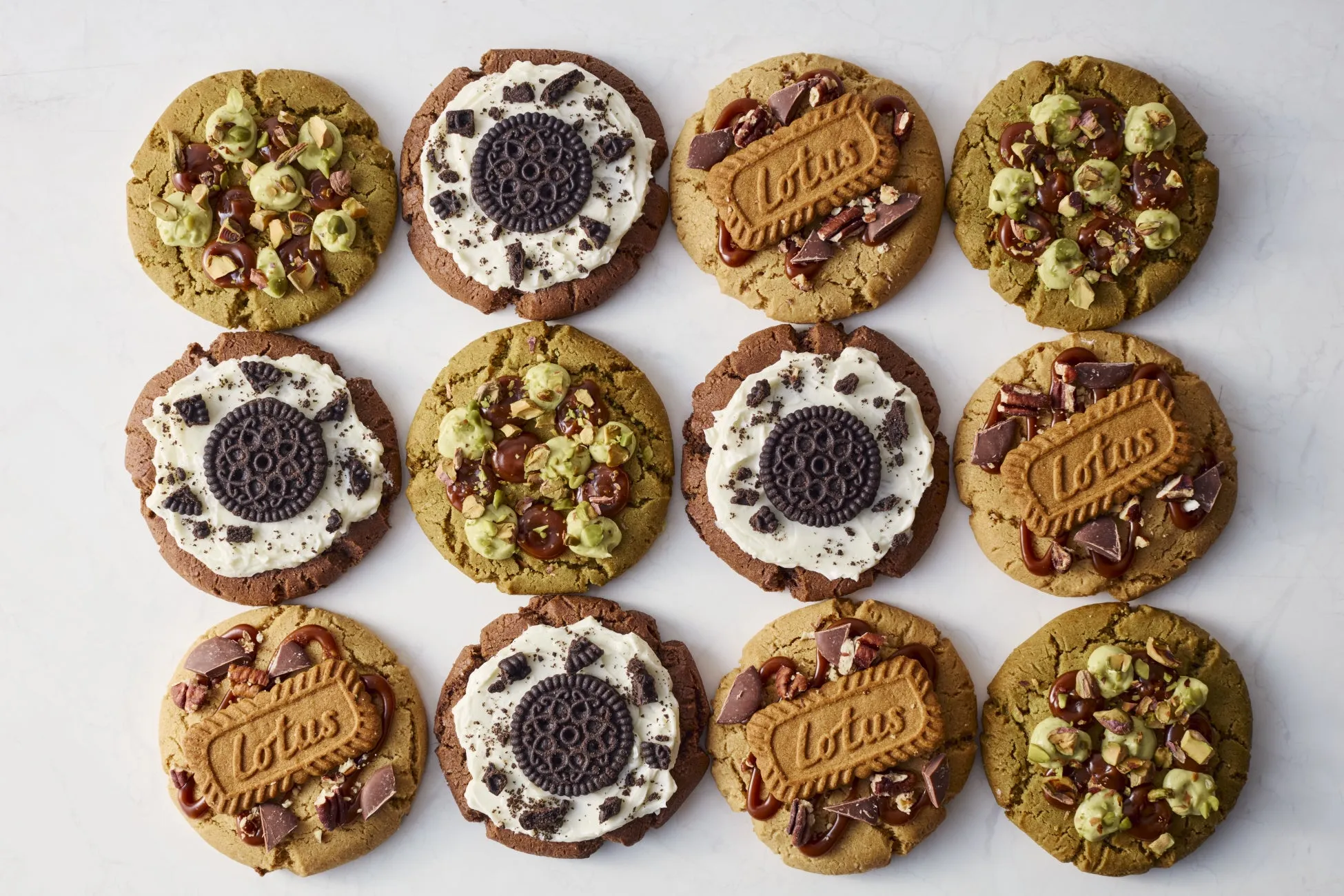 さまざまなトッピングが施されたクッキーの俯瞰写真。上段には、ピスタチオとチョコレートチップをトッピングした緑色のクッキー、オレオと白いクリームをトッピングした茶色のクッキー、ロータスビスケットとチョコレートチップをトッピングした茶色のクッキーが並んでいる。下段にも同様のクッキーが並んでいる。