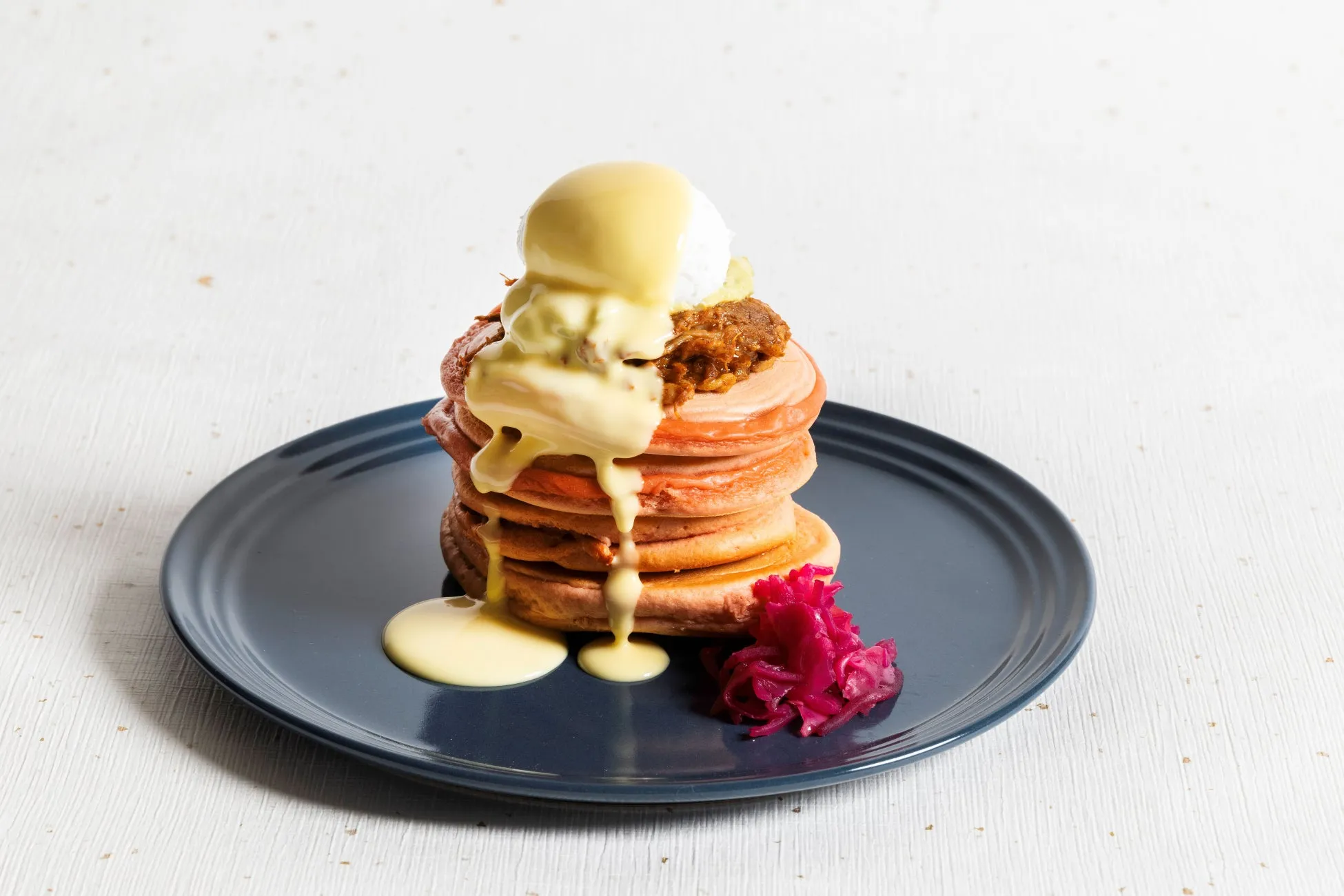 青い皿に、パンケーキが積み重ねられ、ソースがかけられ、付け合わせに紅色の漬物がある料理の写真です。