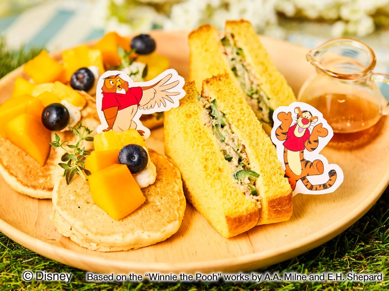 木製のプレートに、パンケーキ、サンドイッチ、マンゴー、ブルーベリー、キャラクターのイラストが配置された、くまのプーさんのテーマの食べ物の写真。