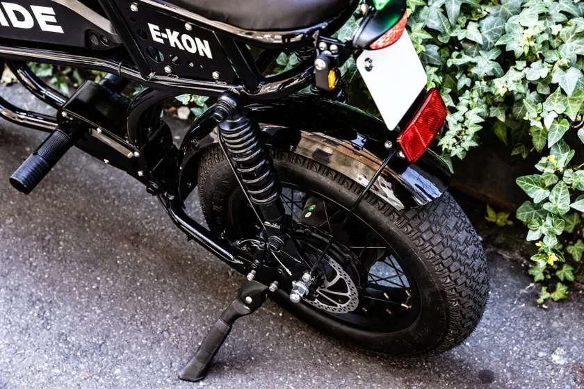 黒い電動バイクの後部のクローズアップ。バイクのタイヤ、サスペンション、ブレーキディスク、ナンバープレート、テールライト、緑の植物が見えます。