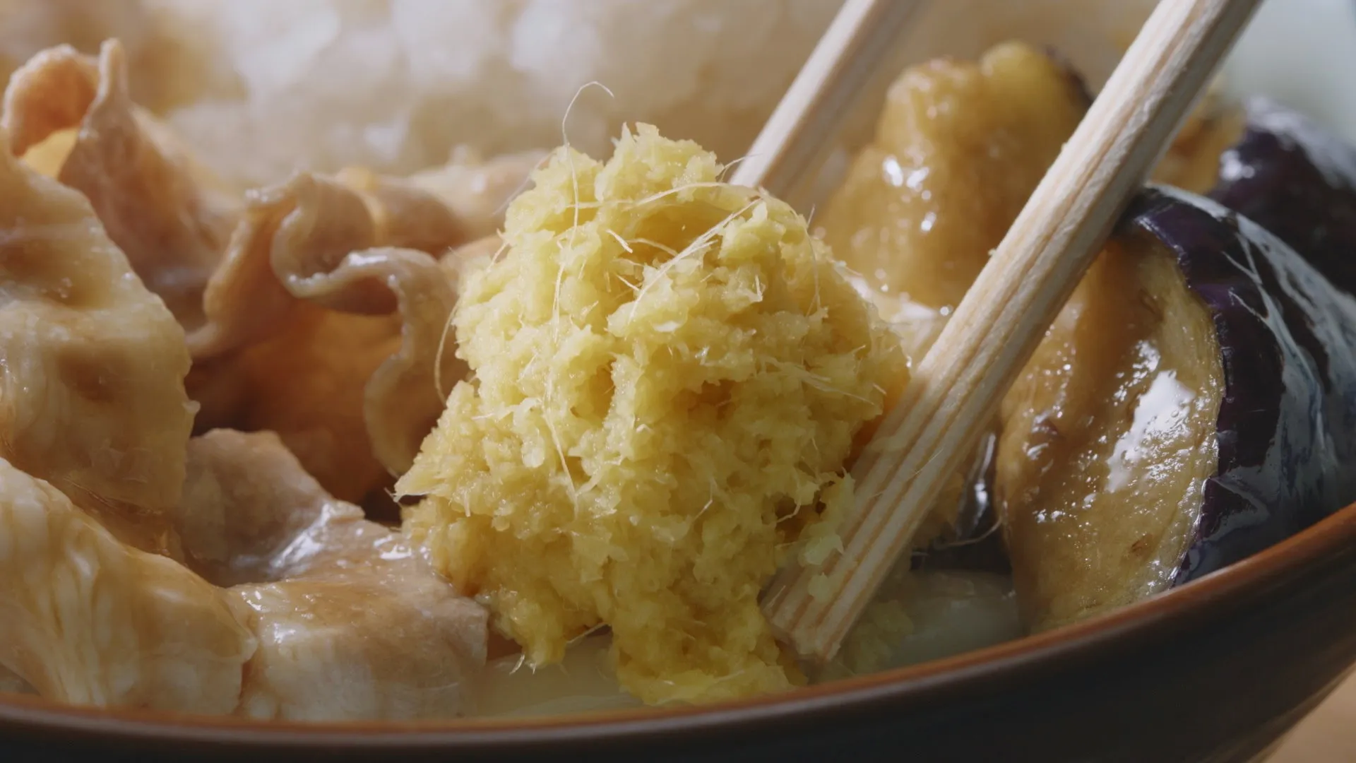 お椀に入った料理の写真です。生姜やナスなどの食材が写っています。箸で生姜を掴もうとしている様子です。