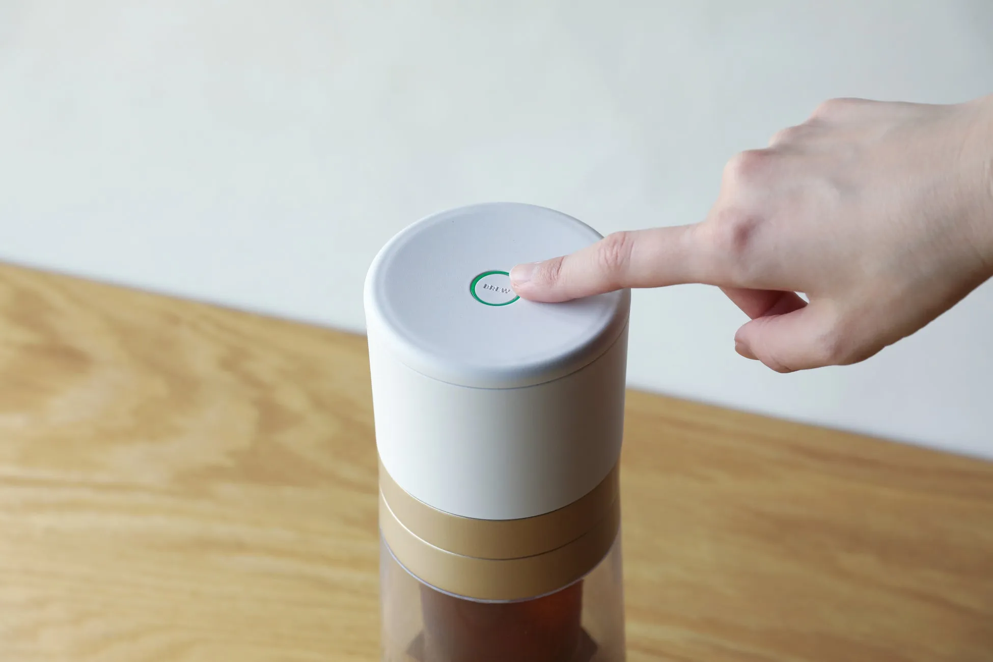 白い円筒形のデバイスの「BREW」と書かれたボタンを指で押している様子が写っています。デバイスは木目調のテーブルの上に置かれており、背景は白です。