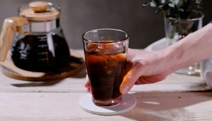 氷が入ったコーヒーが入ったグラスを手に持った様子を捉えた写真です。背景には、コーヒーを淹れるための器具や、花瓶に入った植物が見えます。テーブルは木製で、落ち着いた雰囲気です。