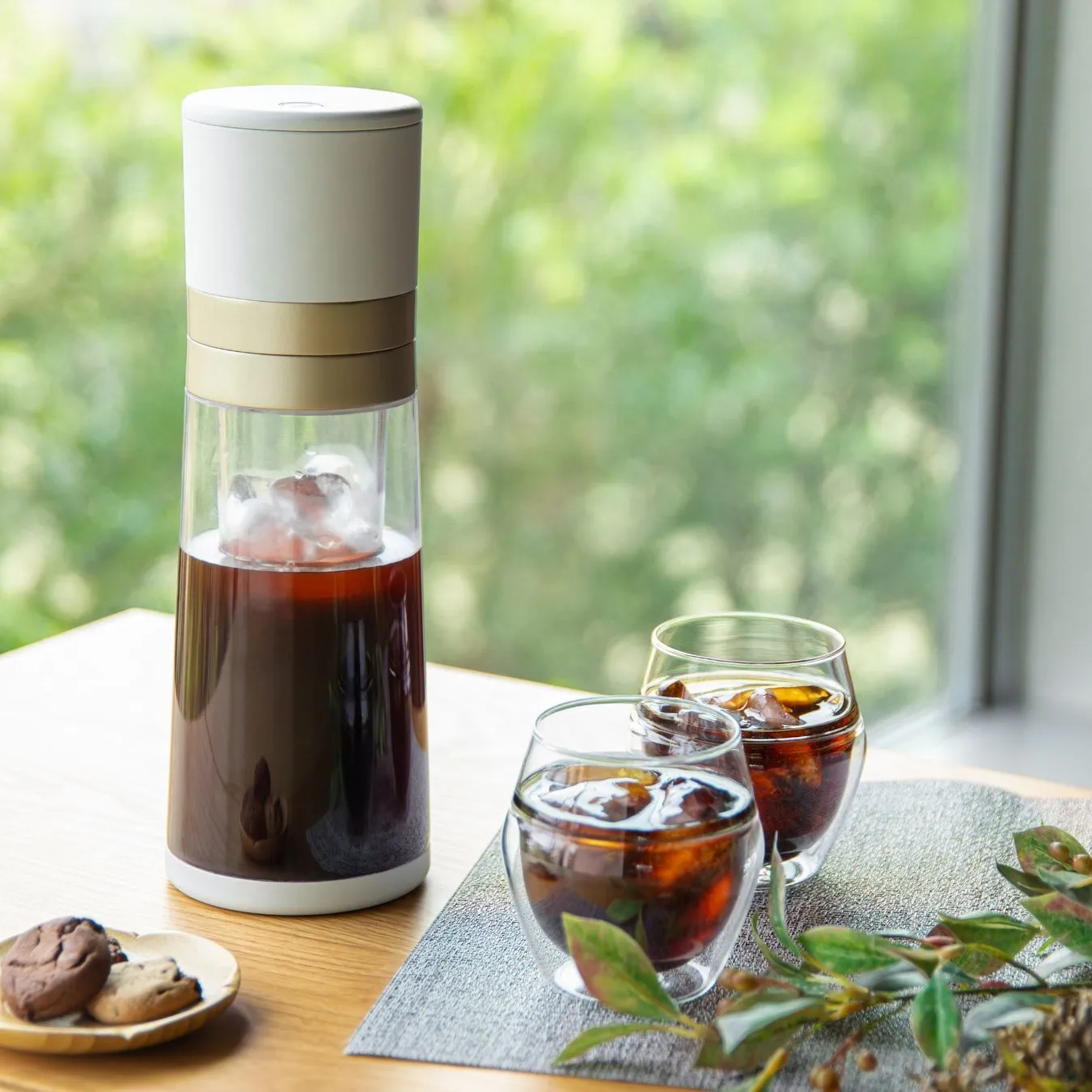 透明な水出しコーヒーメーカーと、氷入りアイスコーヒーが注がれた2つのグラスが木製テーブルに置かれています。隣にはクッキーの小皿と緑の飾りが添えられ、背景の緑が爽やかな、リラックスした家カフェの雰囲気を醸し出しています。